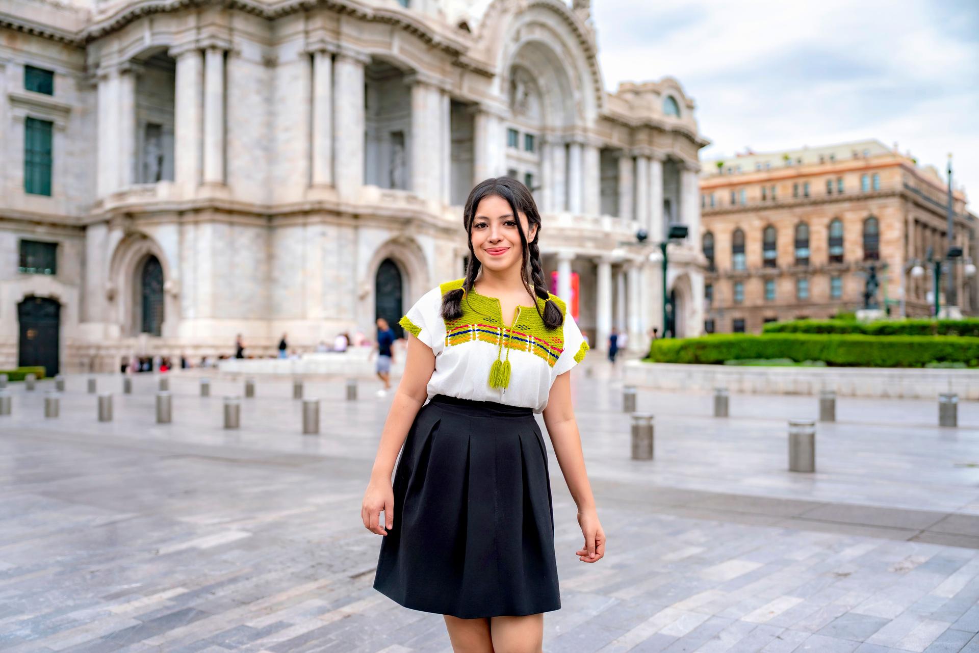 Confident young Latina woman in traditional embroidered top and black skirt smiling in front of Palacio de Bellas Artes, Mexico City